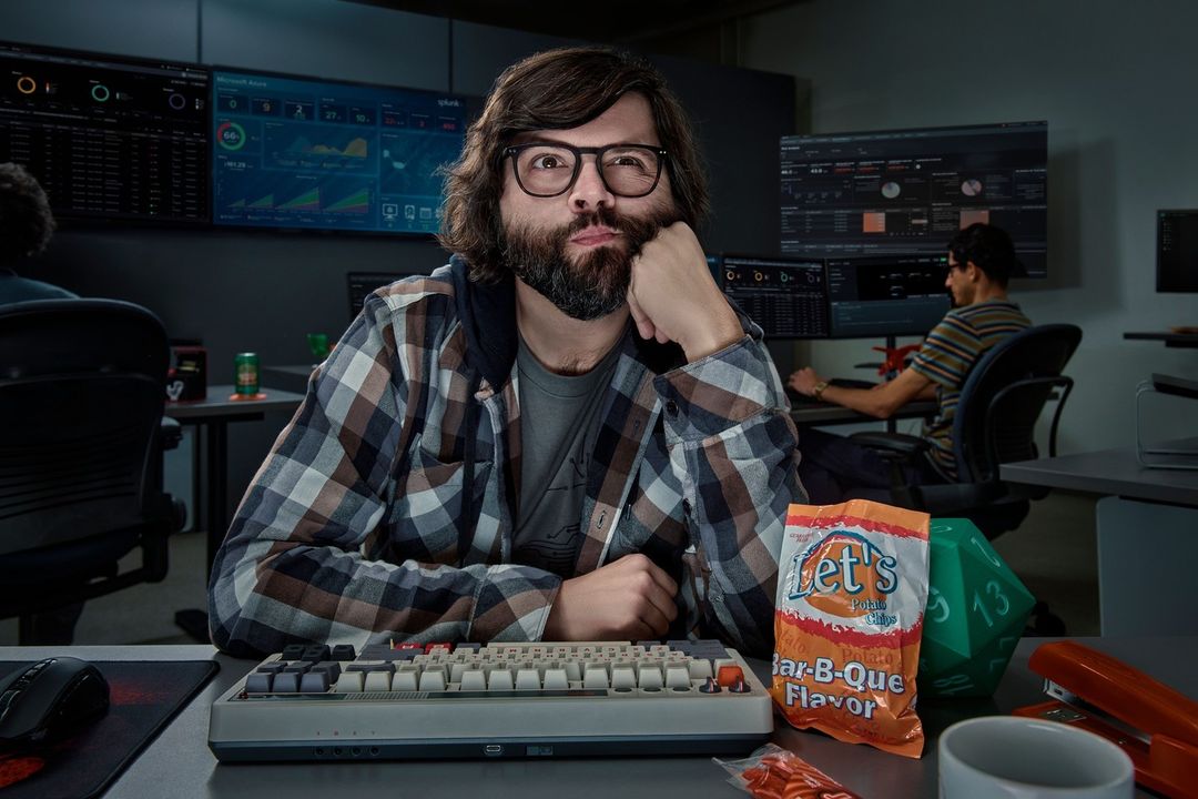 Man with glasses, long hair, and beard leans at a desk with a keyboard, chips, and a multi-monitor workstation in the background.